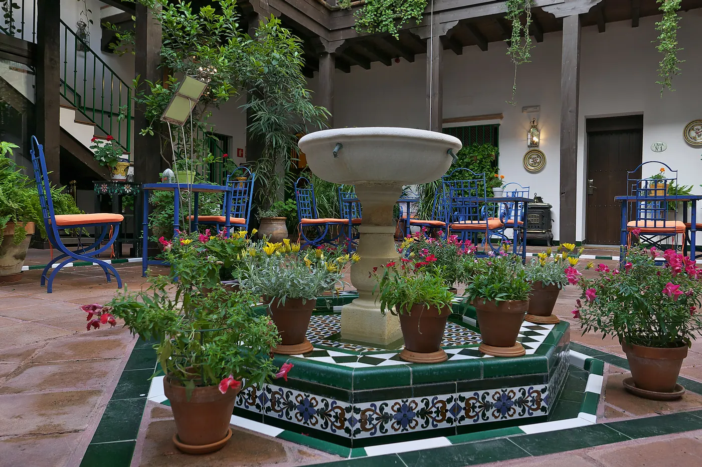 A tiled courtyard fountain ringed by flowerpots, with blue chairs and wooden galleries around it, in the Barrio de Santa Cruz