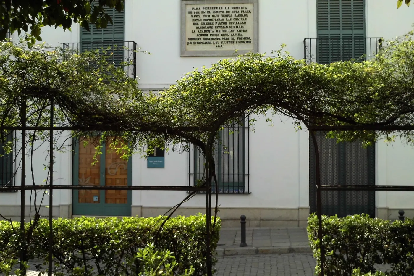 A whitewashed house front with a stone plaque, seen through a trellis arch and clipped hedges in the Plaza de Santa Cruz