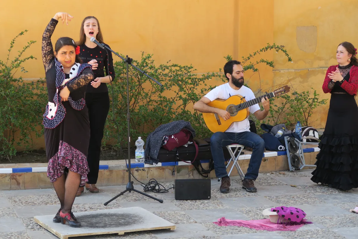 A flamenco dancer in a black polka-dot dress with a raised arm, a singer at a microphone and a seated guitarist in a sunlit yellow courtyard in Seville
